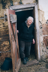 A very old woman with gray hair in old clothes stands on the threshold of a dilapidated house