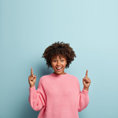 Positive female seller points both fore fingers upwards, has toothy smile, wears casual pink...
