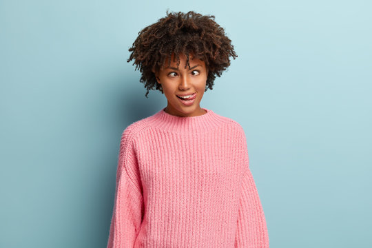 Crazy funny dark skinned woman crosses eyes, makes grimace, sticks out tongue, foolishes indoor, has curly hair and dark skin, isolated over blue background. Human facial expressions, craziness