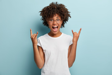 Dark skinned woman shouts from happiness, has widely opened mouth, makes rock n roll gesture, wears casual outfit, says rock this party, feels cool, models over blue background. Body language - Powered by Adobe
