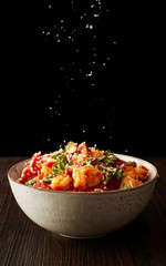 A bowl of Gnocchi pasta on a wood table with salt or cheese being sprinkled on top against a black background