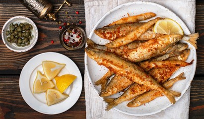Fried smelt in a white plate. Small fish. Capers, lemon, pepper and salt on a wooden table. A...