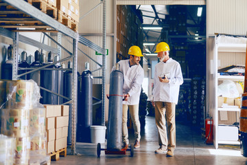 Smiling young Caucasian employee transporting bottle with gas while manager holding tablet and talking to worker. Warehouse interior. - obrazy, fototapety, plakaty