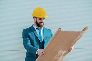 Young attractive successful Caucasian bearded architect with yellow helmet on head and in blue suit looking at blueprints while standing at site.