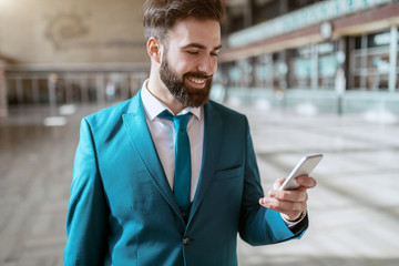 Young attractive bearded smiling businessman in blue suit carrying luggage and using smart phone while standing at train station. Business trip concept.