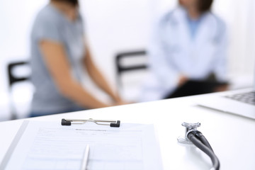 Stethoscope, clipboard with medical form lying on hospital reception desk with laptop computer and busy doctor and patient communicating at the background. Medical tools at doctor working table