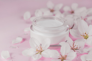 Beauty concept. Jar of face cream surrounded by blossoming spring flowers on pink background close up.
