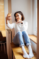 Pretty woman sitting on the windowsill and making selfie on smartphone