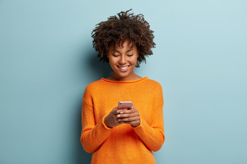Satisfied hipster girl with Afro haircut, types text message on cell phone, enjoys online communication, types feedback, wears orange jumper, isolated on blue studio wall. Technology concept - obrazy, fototapety, plakaty