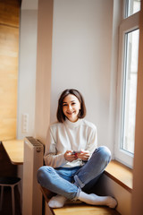 Obraz premium Young woman watches something on the Internet in her mobile phone and smiling while sitting on a windowsill