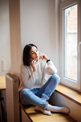 Beautiful young woman sitting on windowsill, laughing and talking on the phone
