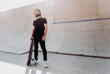 The man dressed in the stylish casual clothes holding skateboard in his hand stands in a skate park next to the slide at the sunny day