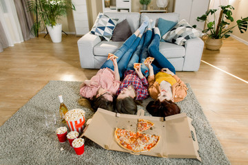 Three Girlfriends in jeans and shirts eating Pizza at home, lying on the floor and put legs on the sofa