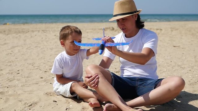 A little cute boy and his mom sit on the sandy shore on a sunny summer day and play with a toy airplane