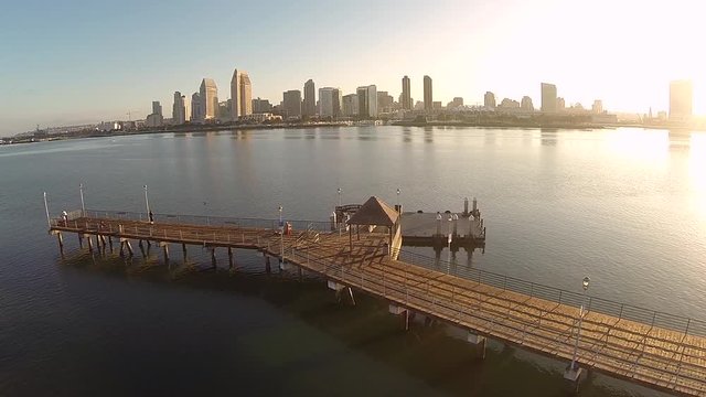 Drone panning shot of wooden boat pier with people fishing in the water channel and the sun shining through the city skyline showing high-rise buildings past the bay in the background. 