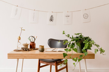 Scandinavian interior of home office space with wooden desk, design chair, forest accessories, avocado plant, plants and  office supplies. Stylish drawings on the white wall. Botanical home decor. 