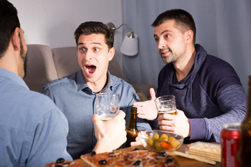 Three cheerful men talking with beer and pizza at home