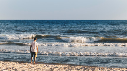 Fototapeta na wymiar Young adult man standing on sand and staring at small waves of sea