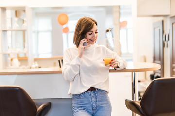 Smiling young woman using mobile phone, holding glass of orange juice at the kitchen