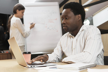 Confident concentrated African man using portable electronic gadget at office desk while his female assistant in background working on presentation, standing at flip chart, reporting about project