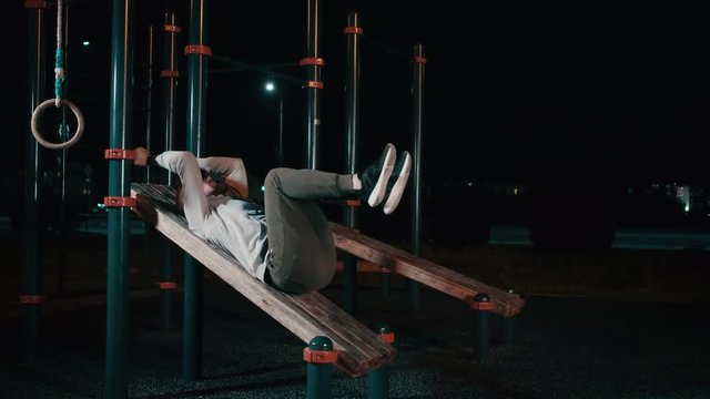 Young sporty man is lifting legs lying on sports equipment in night park