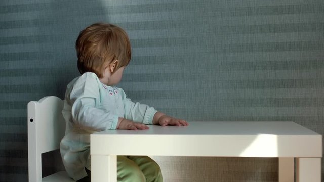 Little cute baby girl is sitting on the chair at the childs small table on the background of beautiful blue wallpaper, turns her head and looks into the camera in slow motion. Suns beam falls on table