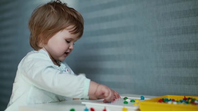 Little Cute Baby Girl Sits At A Childs Table, Smile And Collects A Mosaic Close Up On The Background Of Beautiful Blue Wallpaper In Slow Motion. Child Plays Toys With Enthusiasm