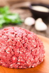 Fresh raw ground or minced beef meat on wooden plate, seasonings, garlic and fresh parsley in the back (Selective Focus, Focus on the front of the meat)