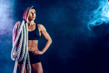 Attractive woman in black sportswear with heavy ropes on her shoulders on dark background. Strength and motivation. Sporty woman working with heavy ropes. Smoke on background.