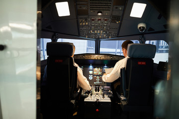 Rear view of serious pilots in white shirts sitting at control panels with knobs and switches while...