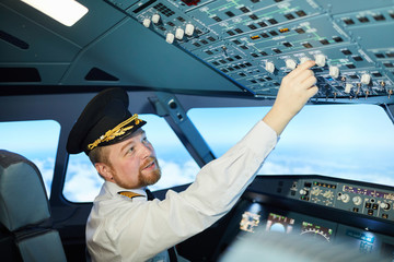 Serious chubby bearded professional pilot in captains cap sitting in armchair and using switches in...