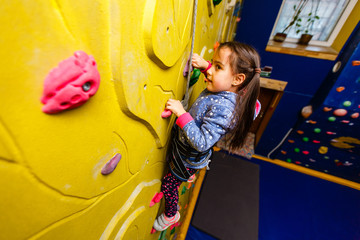 Fototapeta na wymiar Little baby girl with funny hear style climbing vertical wall and man belaying her from below
