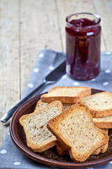 Toasted cereal bread slices on grey plate and jar with homemade cherry jam closeup on linen napkin on rustic wooden table background.
