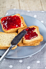 Fresh toasted cereal bread slices with homemade cherry jam and knife on grey plate closeup on grey linen napkin background.