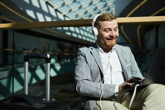 Cheerful excited chubby young man with beard sitting in waiting area of airport and using laptop while browsing internet and listening to music in headphones