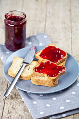 Toasted cereal bread slices on grey plate and jar with homemade cherry jam closeup on linen napkin on rustic wooden table background.