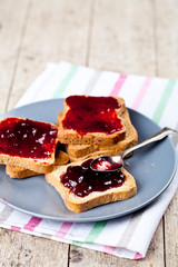 Fresh toasted cereal bread slices with homemade cherry jam and spoon on ceramic plate closeup on linen napkin on rustic wooden table background.