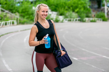 Fitness lifestyle. Young woman holding a water bottle and a bag on her shoulder after a workout. Workout at the stadium. Healthy life concept