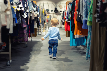 Cute caucasian blond toddler boy walking alone at clothes retail store between rack with hangers....
