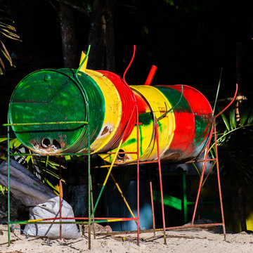 Jamaican jerk pan/ grill in Rasta colors in Jamaica. Used to cook jerk fish, chicken, pork meals outdoors. It is a metal drum cut down the middle, placed sideways on metal stand, filled with coal.