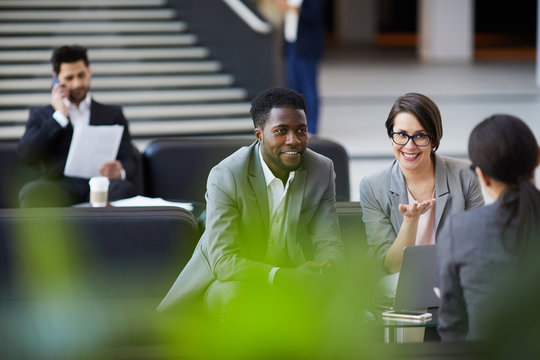 Cheerful positive multi-ethnic business employees in formalwear sitting on sofas in lobby and planning strategy for project implementation