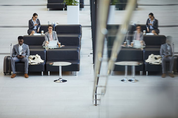 Serious multi-ethnic business people sitting on sofas and using modern devices while waiting for boarding in airport business class lounge, reflection effect