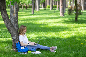 Adorable freelance woman in white t-shirt and jeans sit on the grass under tree in park with laptop on her legs. She smile, listening music and typing text on computer. Coffee, notebooks near