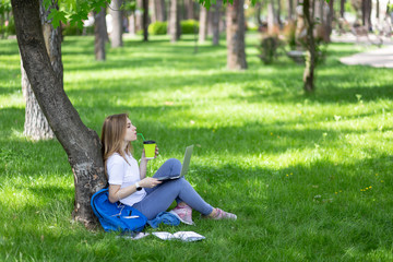 Naklejka na ściany i meble Adorable woman in white t-shirt and jeans drink coffee sitting on the grass under tree with laptop on her legs. She listening music with closed eyes. Notebooks, dark-blue backpack, phone near. Copy sp