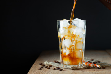 Ice coffee in a tall glass with cream poured over, ice cubes and beans on a old rustic wooden table. Cold summer drink with tubes on a black background with copy space