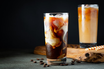 Ice coffee in a tall glass with cream poured over, ice cubes and beans on a old rustic wooden table. Cold summer drink with tubes on a black background with copy space - obrazy, fototapety, plakaty