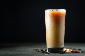 Ice coffee in a tall glass with cream poured over, ice cubes and beans on a old rustic wooden...