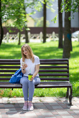 Adorable young blonde woman in white t-shirt and blue jeans sits on the bench with smartphone and cup of coffee in her hands. She look at smartphone in her hand and listening music via wired earphones