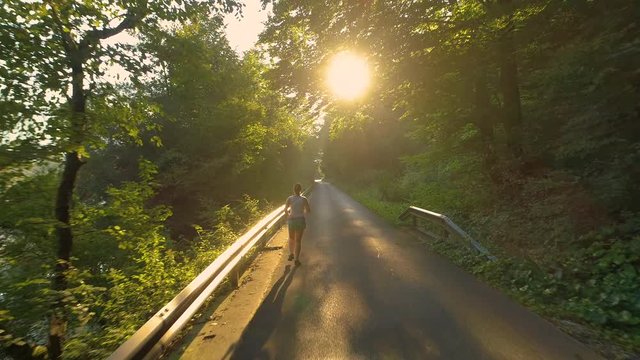 AERIAL, LENS FLARE: Unrecognizable young woman runs down an empty asphalt road leading through the calm woods. Flying behind unrecognizable fit girl exercising in nature on a beautiful spring morning.