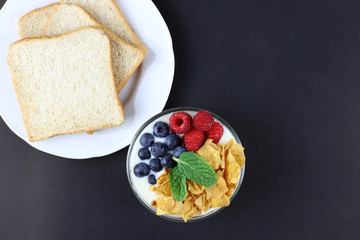 Sliced ​​bread on white plate and bowl of yogurt with raspberry and blueberry and cereals on black background